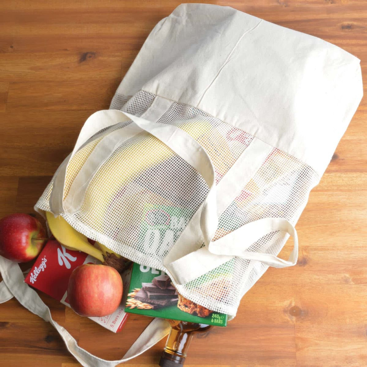 A calico and mesh tote bag filled with fresh apples, a box of cereals, a pack of cookies, and a bunch of bananas